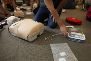 A CPR dummy with medical equipment attached to it. Someone is kneeling by it. 